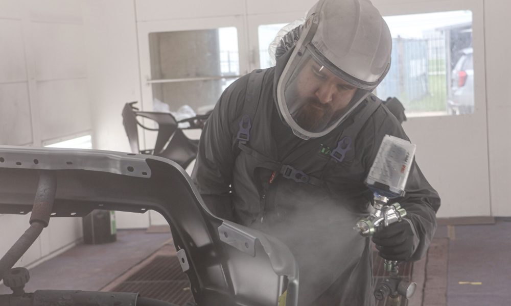 A Core Collision paint technician applies automotive paint to a vehicle panel inside the paint booth in Georgetown, TX while wearing protective equipment.